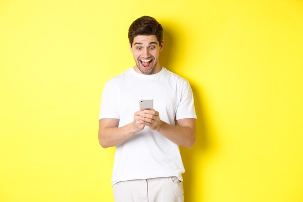Hombre sorprendido leyendo un mensaje de texto en el teléfono móvil, mirando sorprendido y feliz en la pantalla del teléfono inteligente, de pie sobre fondo amarillo