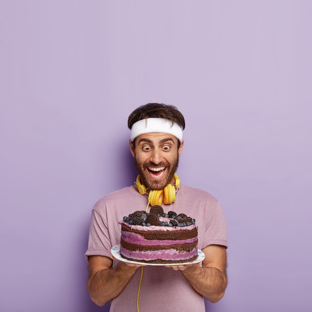 El hombre sorprendido y lleno de alegría mira un pastel delicioso, siente la tentación, tiene hambre después del entrenamiento activo, usa una camiseta informal, lleva auriculares