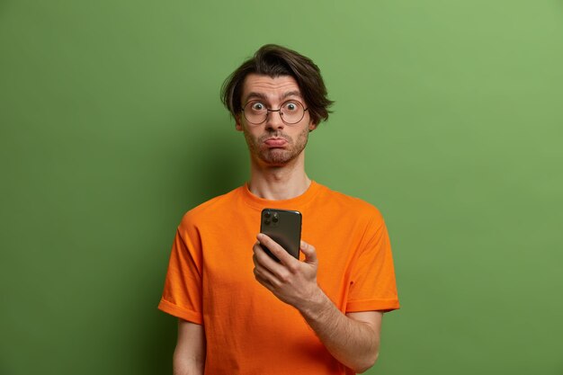 Hombre sorprendido ofendido con peinado moderno, cara infeliz porque no puede hacer un pedido en línea, sostiene un teléfono inteligente moderno, vestido con una camiseta naranja, aislado en una pared verde. Concepto de tecnología