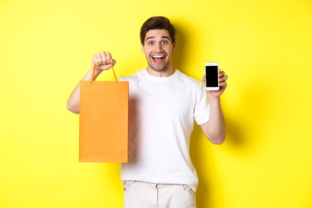 Hombre sorprendido sosteniendo la bolsa de compras y mostrando la pantalla del teléfono inteligente, concepto de banca móvil y logros de aplicaciones, fondo amarillo.