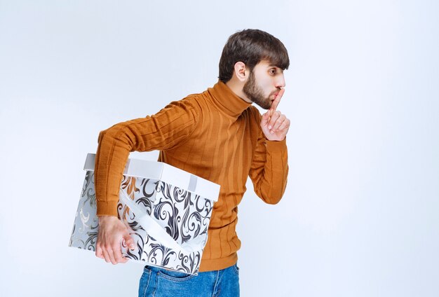 Hombre sosteniendo una caja de regalo blanca con patrones azules y pidiendo silencio o dudando sobre algo.