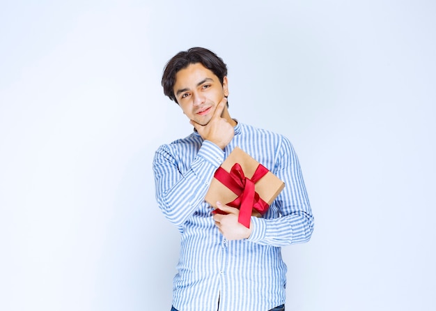 Hombre sosteniendo una caja de regalo de cartón con cinta roja y dudando o avergonzado. Foto de alta calidad