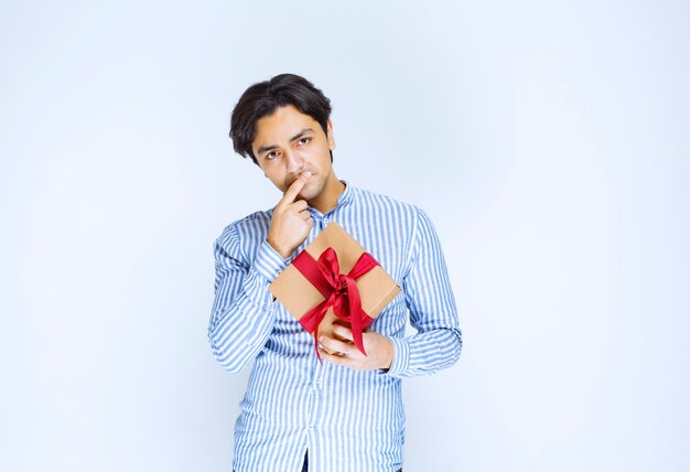 Hombre sosteniendo una caja de regalo de cartón con cinta roja y dudando o avergonzado. Foto de alta calidad