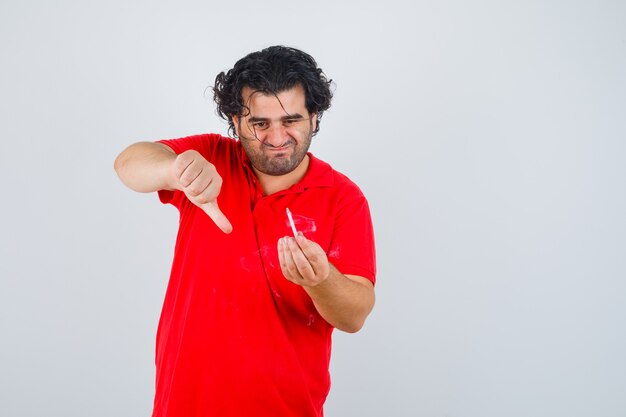 Hombre sosteniendo un cigarrillo, mostrando el pulgar hacia abajo en una camiseta roja y mirando enfocado, vista frontal.