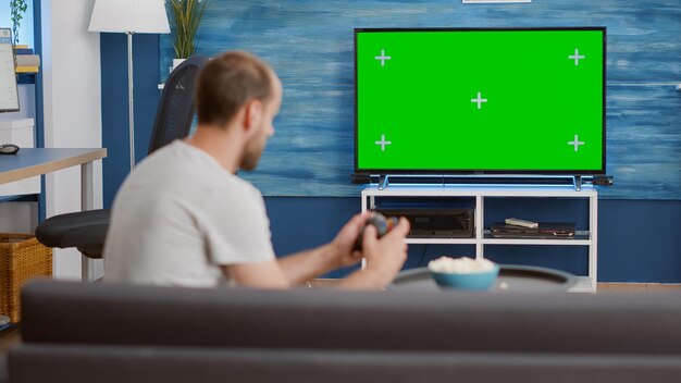 Hombre sosteniendo un controlador inalámbrico jugando videojuegos de consola en una televisión de pantalla verde mientras se sienta en un sofá en una sala de estar moderna. Jugador relajándose en el sofá disfrutando de los juegos en línea en la pantalla croma key.