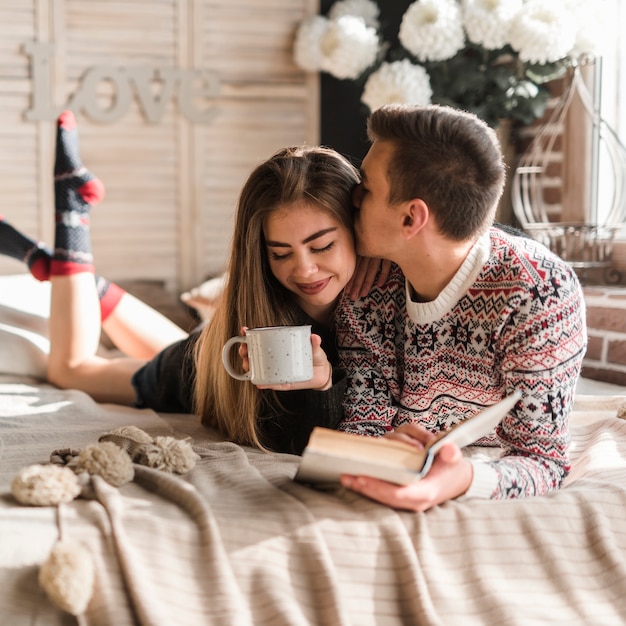 Hombre sosteniendo un libro en la mano besando a su novia sosteniendo una taza de café acostada en la cama