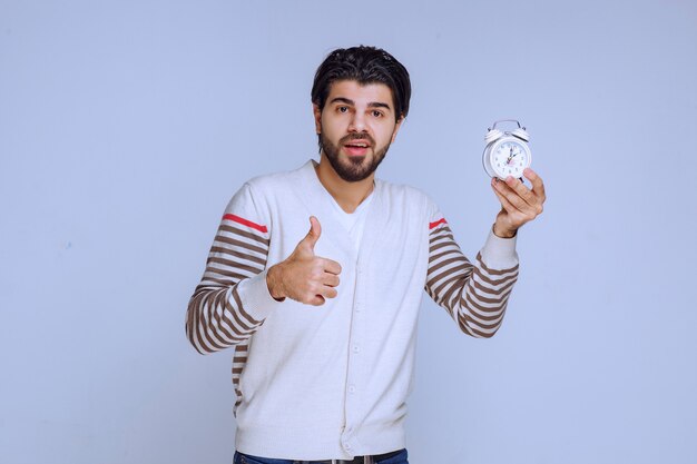 Hombre sosteniendo un reloj despertador y haciendo pulgar hacia arriba.