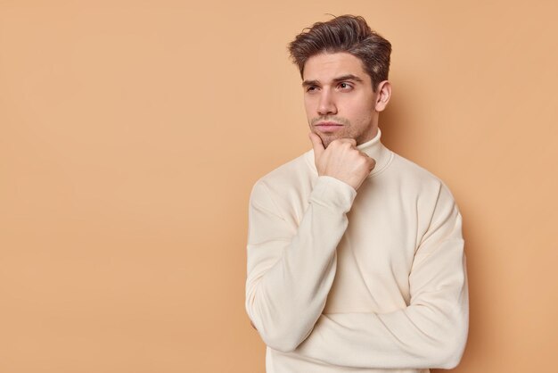 El hombre sostiene la barbilla mira hacia otro lado pensativamente concentrado considera los planes futuros vestido con un cuello alto informal aislado en beige con espacio para copiar el texto