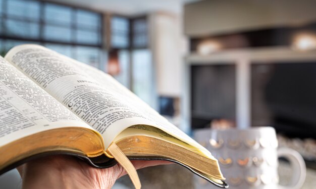 Un hombre sostiene una Biblia en el contexto de la sala de estar. Leer un libro en un ambiente acogedor. De cerca.