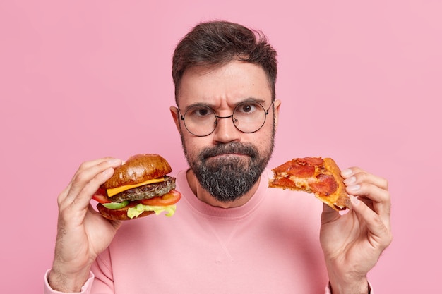 El hombre sostiene la hamburguesa y la pizza presiona los labios lleva gafas redondas puente