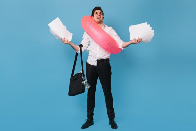 El hombre sostiene un montón de papeles de trabajo sobre fondo azul El tipo con ropa de oficina sostiene una máscara de buceo de bolsa negra y un anillo de goma rosa