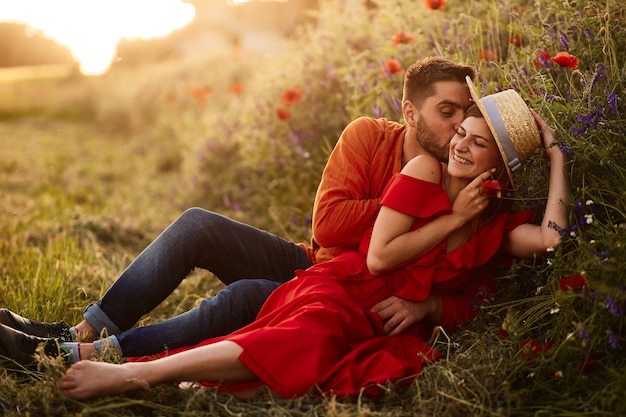 Foto gratuita el hombre sostiene a su mujer tierna sentada con ella en el césped verde con amapolas rojas