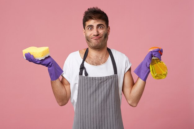 Hombre sucio con peinado elegante y cerdas con esponja y detergente con camiseta blanca informal, delantal y guantes cansados