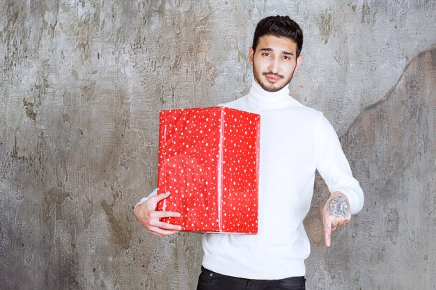 Foto gratuita hombre de suéter blanco sosteniendo una caja de regalo roja con puntos blancos e invitando a alguien a su lado