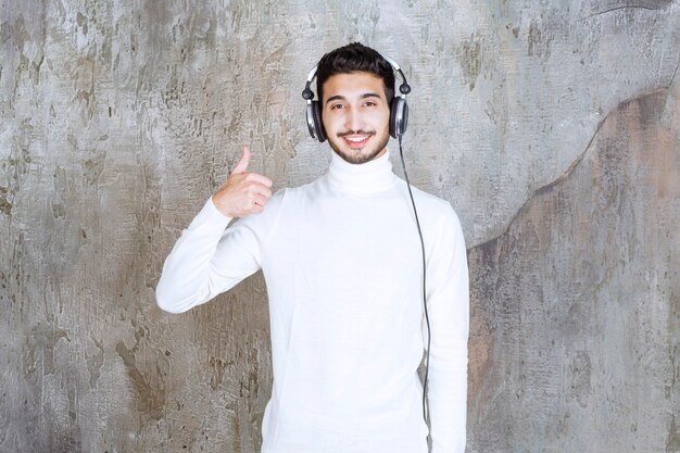 Hombre de suéter blanco usando audífonos y escuchando música y mostrando un signo de mano positivo.