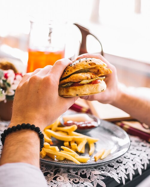 Hombre sujetando una hamburguesa de pollo servida con papas fritas y salsa