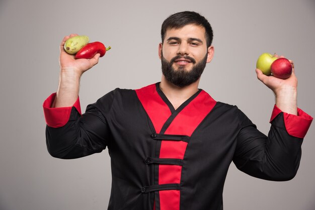 Hombre sujetando pimiento rojo, manzanas y calabacín.