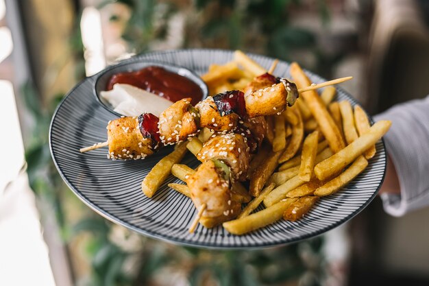 Hombre sujetando un plato de pollo kebab adornado con sésamo servido con papas fritas