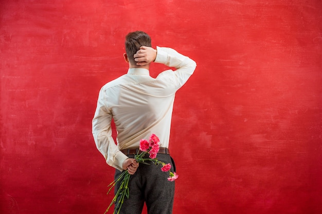 Foto gratuita hombre sujetando ramo de claveles detrás de la espalda sobre fondo rojo studio
