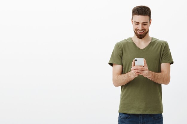 Hombre sujetando un teléfono móvil mirando la pantalla del dispositivo con una sonrisa de satisfacción, enviando mensajes o viendo videos divertidos y divertidos en línea sobre una pared blanca