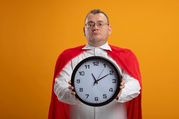 Hombre de superhéroe adulto en capa roja con gafas mirando al frente estirando el reloj hacia el frente aislado en la pared naranja