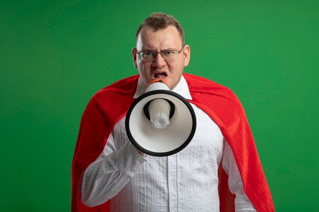 Hombre de superhéroe adulto con el ceño fruncido en capa roja con gafas mirando al frente hablando por altavoz aislado en la pared verde