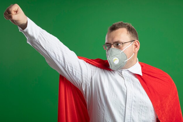 Hombre de superhéroe adulto confiado en capa roja con gafas y máscara protectora mirando al lado levantando el puño aislado en la pared verde