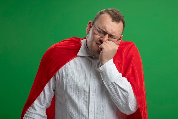 Hombre de superhéroe adulto dolorido en capa roja con gafas poniendo la mano en la mejilla que sufre de dolor de muelas con los ojos cerrados aislado en la pared verde