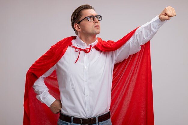 Hombre de superhéroe en capa roja y gafas mirando a un lado con cara seria haciendo gesto ganador con la mano lista para ayudar a pararse sobre la pared blanca