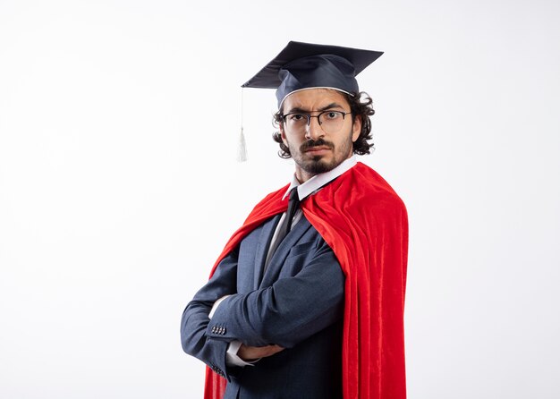 Hombre de superhéroe caucásico joven confiado en gafas ópticas con traje con capa roja y gorro de graduación se encuentra de lado con los brazos cruzados mirando con espacio de copia