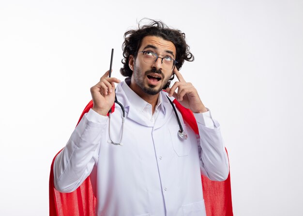 Hombre de superhéroe caucásico joven sorprendido con gafas ópticas con uniforme médico con capa roja y con estetoscopio alrededor del cuello mira y apunta con lápiz con espacio de copia