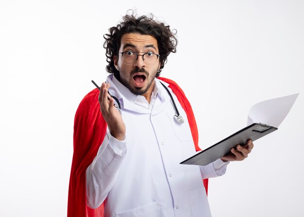 Hombre de superhéroe caucásico joven sorprendido con gafas ópticas con uniforme de médico con capa roja y con estetoscopio alrededor del cuello sostiene lápiz y portapapeles con espacio de copia