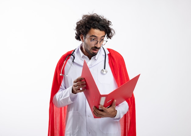 Hombre de superhéroe caucásico joven sorprendido con gafas ópticas con uniforme médico con manto rojo y con estetoscopio alrededor del cuello sostiene y mira la carpeta de archivos con espacio de copia