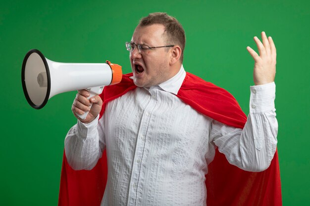 Hombre de superhéroe eslavo adulto con el ceño fruncido en capa roja con gafas gritando en altavoz manteniendo la mano en el aire mirando al lado aislado en la pared verde