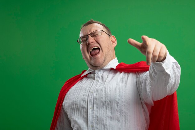 Hombre de superhéroe eslavo adulto confiado en capa roja con gafas apuntando con los ojos cerrados aislado en la pared verde con espacio de copia