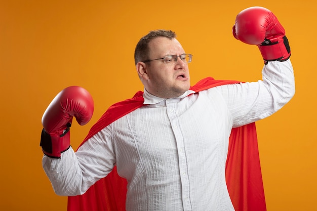 Foto gratuita hombre de superhéroe eslavo adulto confiado en capa roja con gafas y guantes de caja haciendo un gesto fuerte mirando al lado aislado en la pared naranja