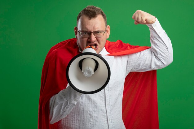 Hombre de superhéroe eslavo adulto confiado en capa roja con gafas haciendo gesto fuerte hablando por altavoz aislado en pared verde