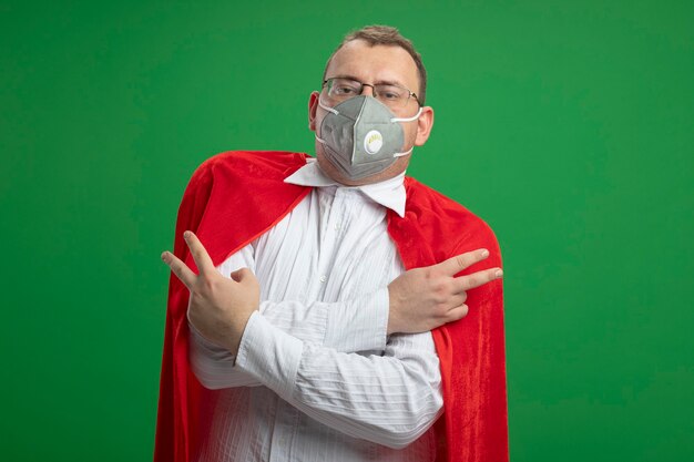 Hombre de superhéroe eslavo adulto confiado en capa roja con gafas y máscara protectora manteniendo las manos cruzadas haciendo el signo de la paz aislado en la pared verde con espacio de copia