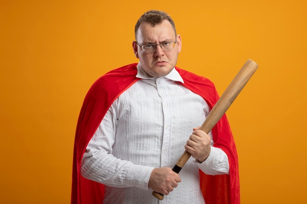 Foto gratuita hombre de superhéroe eslavo adulto frunciendo el ceño en capa roja con gafas sosteniendo un bate de béisbol aislado en la pared naranja con espacio de copia