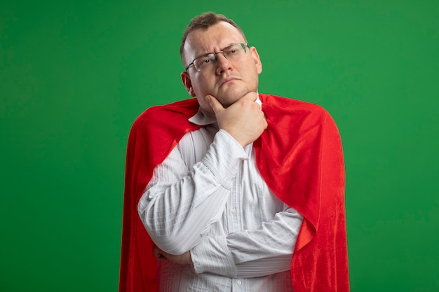 Foto gratuita hombre de superhéroe eslavo adulto pensativo en capa roja con gafas tocando la barbilla mirando al lado aislado sobre fondo verde con espacio de copia