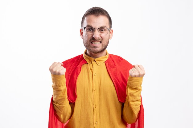 Hombre de superhéroe molesto en gafas ópticas con capa roja mantiene los puños aislados en la pared blanca