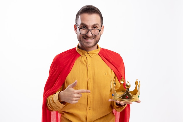 Foto gratuita hombre de superhéroe sonriente en gafas ópticas con manto rojo sostiene y apunta a la corona aislada en la pared blanca