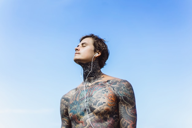 hombre tatuado con auriculares contra el cielo azul en el océano
