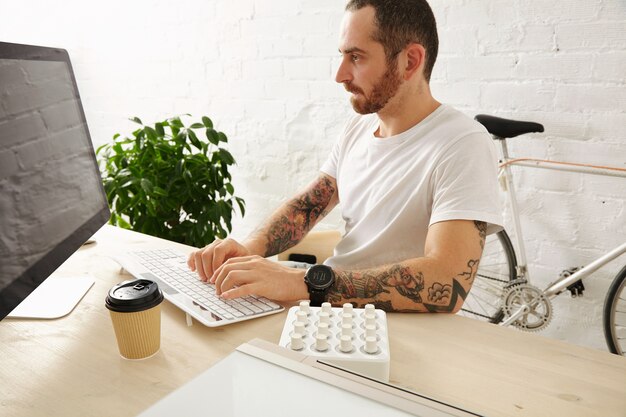 Hombre tatuado con barba en camiseta blanca en blanco trabaja en su computadora en casa, vista lateral, horario de verano