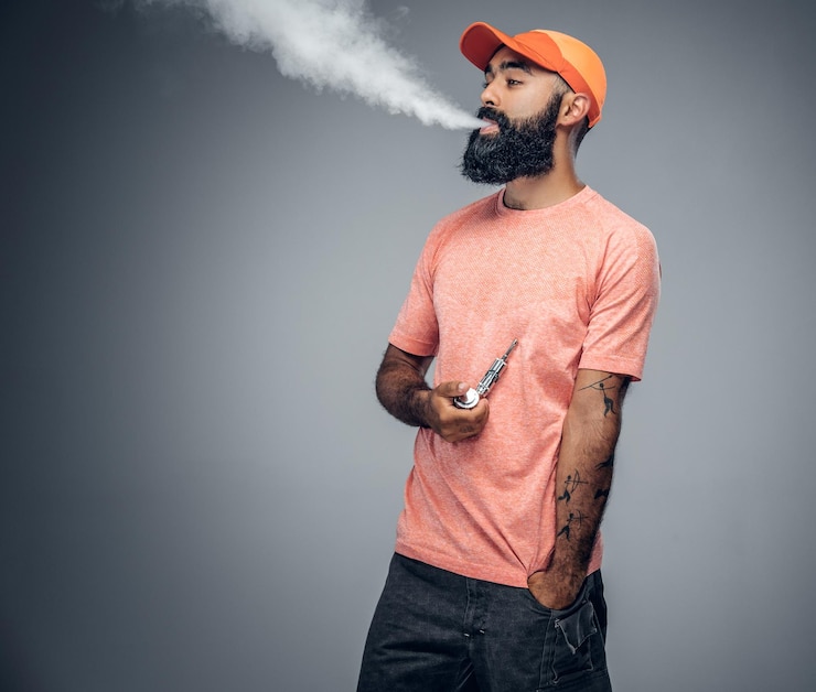 Foto gratuito hombre tatuado barbudo alegre fuma cigarrillo electrónico aislado sobre fondo gris.