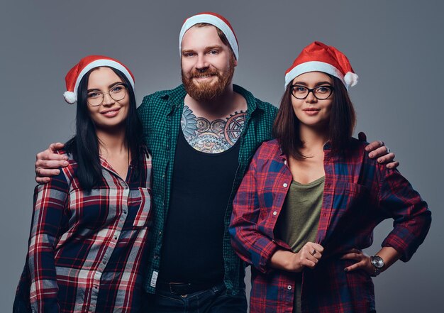 Un hombre tatuado y barbudo y dos mujeres morenas vestidas con ropa navideña posando sobre un fondo gris.