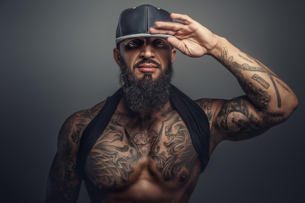 Hombre tatuado brutal con gorra negra posando en el estudio. Aislado sobre fondo gris.