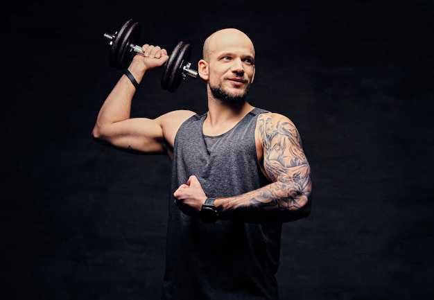 Hombre tatuado con cabeza rapada atlética haciendo ejercicios de hombro con mancuernas.