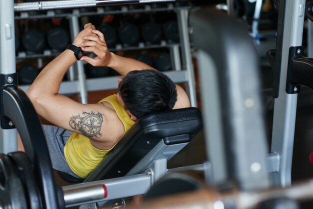 Hombre tatuado recostado en el banco en el gimnasio y mirando el reloj inteligente