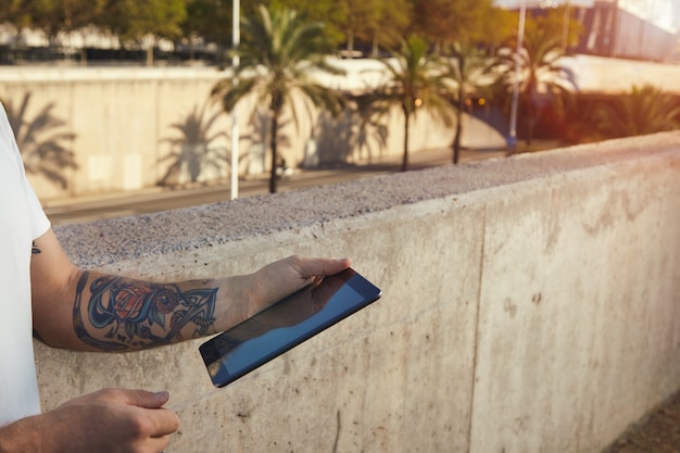 Hombre tatuado sosteniendo una tableta negra de pie junto a un muro de hormigón gris en el paisaje de la ciudad con palmeras
